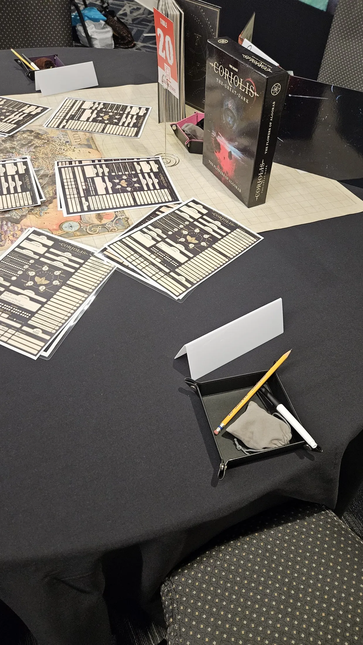 A convention game table at Origins, Table 20, set for Coriolis: The Great Dark. Explorer sheets are fanned across an illustrated Ship City map, with the Flowers of Algorab box, dice trays, and pencils.