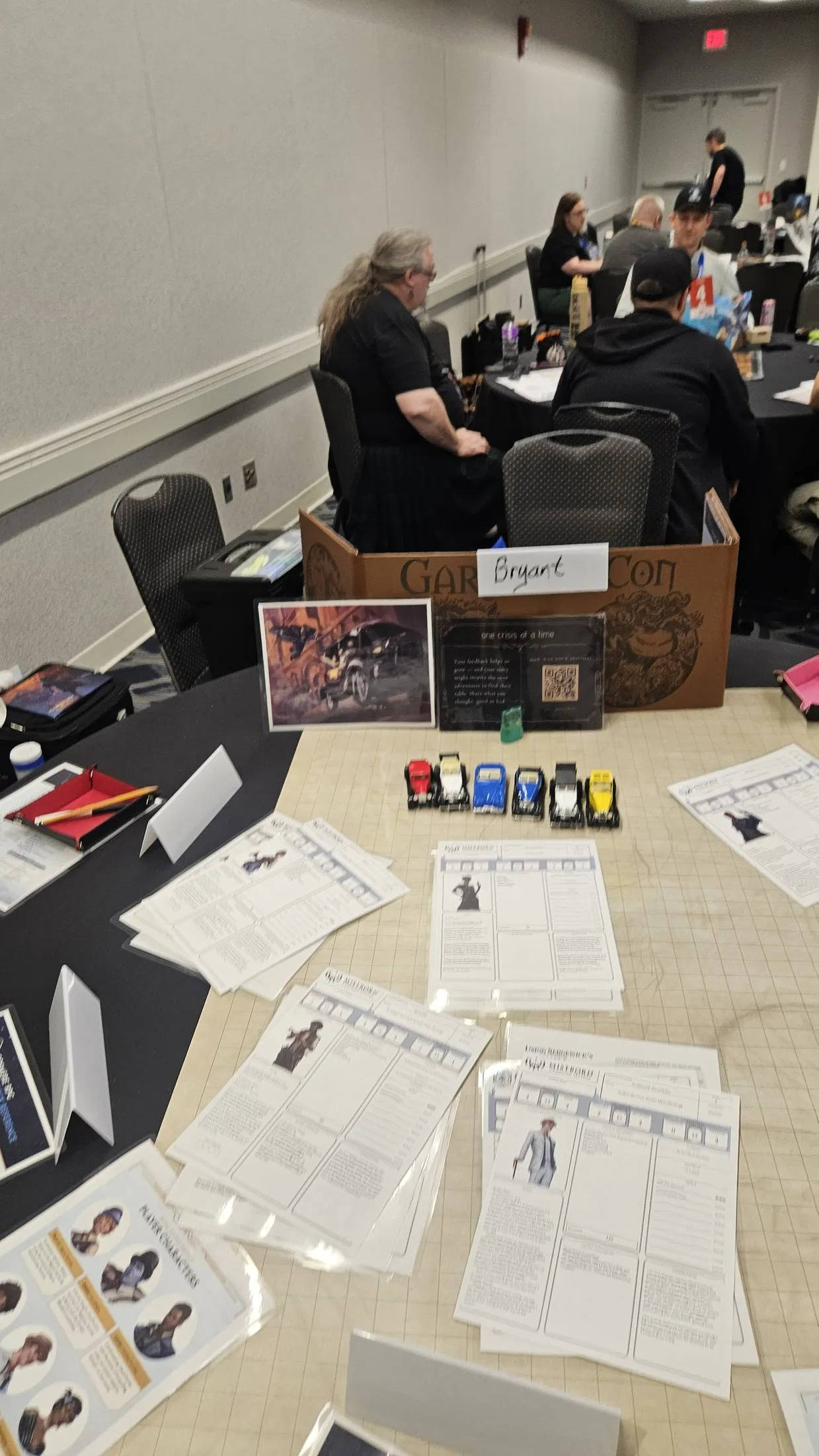 A convention game table set for Pedal to the Metals, with a row of classic toy race cars, printed Mistborn character sheets, dice trays, and an OCAAT table card.